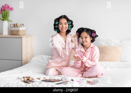 Sorridente giovane cinese femmina fare acconciatura con curlers per ragazza giovane siede sul letto con cosmetici in camera da letto Foto Stock