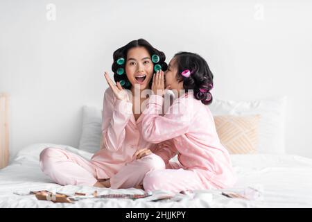 Scioccata giovane donna cinese ascoltare gossip da ragazza teen con curlers, sedersi a letto con cosmetici in camera da letto Foto Stock