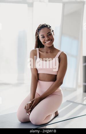 Sorridente giovane afroamericana che indossa abiti sportivi, seduta su materassino fitness, prendendo pausa da casa allenamento Foto Stock