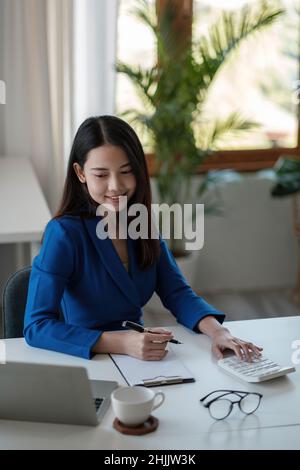 Ritrarre la giovane donna d'affari asiatica seduta alla scrivania e prendere appunti utilizzando il computer portatile e la calcolatrice in ufficio. Foto Stock