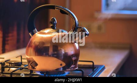 Primo piano di teiera d'argento bollente su stufa. Concetto. bollitore bollente sul piano cottura in bella cucina interna sera. Dal kettl fuoriesce del vapore Foto Stock