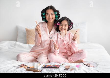 Felice millennial giapponese femmina e adolescente ragazza con curlers tenere le spazzole sedersi a letto con cosmetici Foto Stock