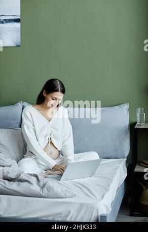 Giovane donna incinta che lavora online su un computer portatile mentre si siede sul letto in camera da letto Foto Stock