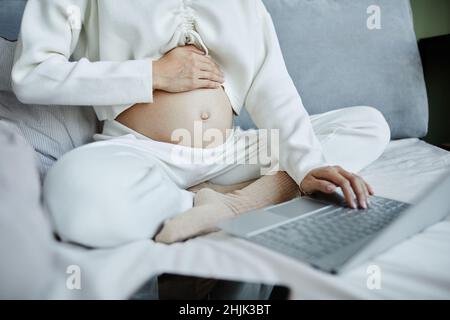 Primo piano di una donna incinta che tocca il ventre mentre si siede sul letto e lavora online sul suo laptop Foto Stock