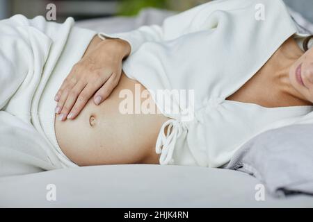Primo piano di una giovane donna incinta che tocca il ventre adagiata sul letto in camera da letto Foto Stock