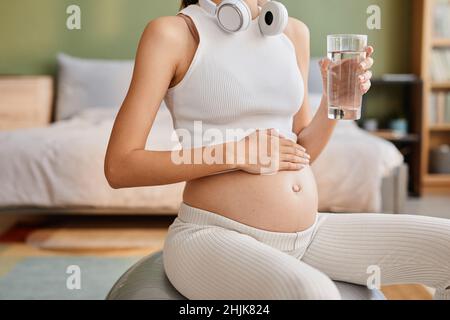 Primo piano di una donna incinta seduta sulla palla fitness e acqua potabile durante il suo allenamento a casa Foto Stock