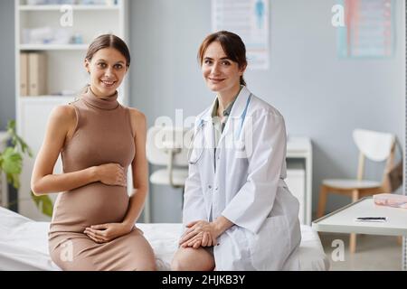 Ritratto della donna incinta e del suo ginecologo sorridendo insieme alla macchina fotografica mentre si siede sul divano all'ufficio del medico Foto Stock