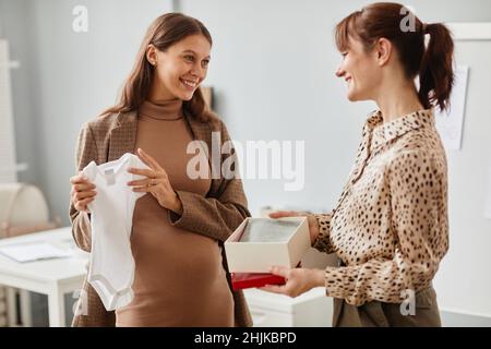 Felice giovane donna incinta che guarda i vestiti del bambino dando dalla sua collega, che parlano in piedi in ufficio Foto Stock