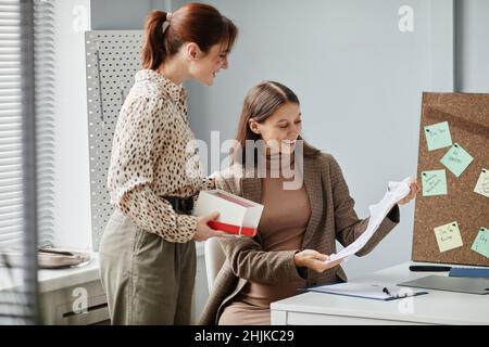 Donna incinta felice seduta sul suo posto di lavoro e disimballare i vestiti dando dalla sua collega per il bambino Foto Stock