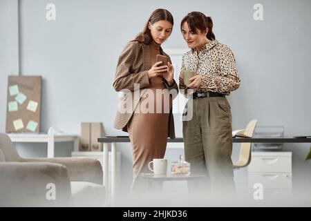 Donna d'affari incinta che mostra le foto sul telefono cellulare al suo collega mentre bevono caffè insieme in ufficio Foto Stock