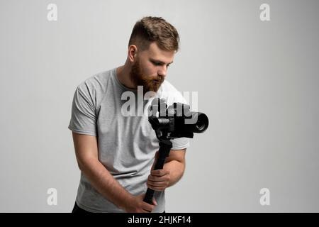 Filmmaking, hobby e concetto di creatività. Videografo maschile professionale che realizza video utilizzando la moderna fotocamera reflex digitale su una parete bianca con sospensione cardanica a 3 assi. Foto Stock
