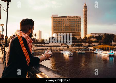 Cairo Egitto Dicembre 2021 uomo appoggiato sulla recinzione di un ponte che conduce alla zona di Zamalek al cairo egitto, cairo torre che si alza in lontananza. Fiume Nilo Foto Stock