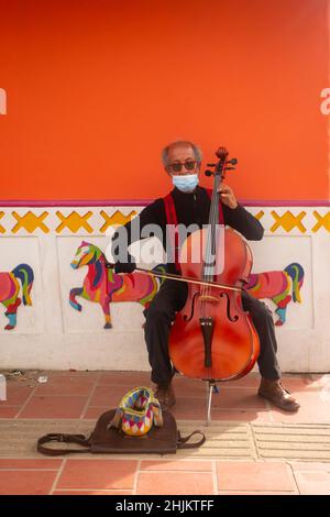 Guatape, Antioquia, Colombia - Dicembre 8 2021: Vecchio uomo marrone vestito in nero con i sospenditori rossi e gli occhiali da sole sta giocando il Cello su una strada Foto Stock