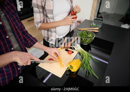 Primo piano delle mani che tritano le verdure su un tagliere e vista ritagliata a una donna incinta che tiene il rametto con pomodori freschi mentre si diverte a cucinare Foto Stock