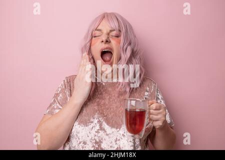 Giovane donna che beve il tè al mattino. Sonnolenta stanca capelli rosa femmina copre bocca e yawns isolato su sfondo rosa. Foto Stock