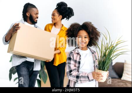 Concetto di rilocazione. Ragazza afroamericana piccola felice che tiene flowerpot, genitori che tengono scatole di cartone, famiglia nera felice che si muove in nuova casa, comprando la loro prima casa, investita nella propria casa Foto Stock