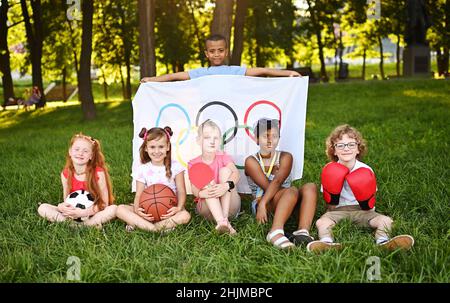 DONETSK, UCRAINA - 10 LUGLIO 2021. I bambini di diverse nazionalità con attrezzature sportive in mano tengono una grande bandiera con simboli olimpici Foto Stock