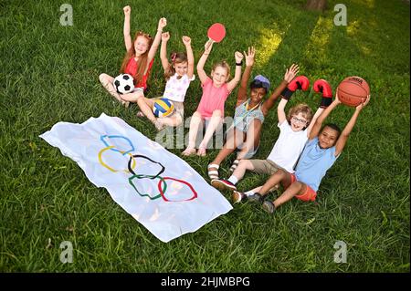 DONETSK, UCRAINA - 10 LUGLIO 2021. I bambini di diverse nazionalità con attrezzature sportive nelle loro mani sono seduti sull'erba contro Foto Stock