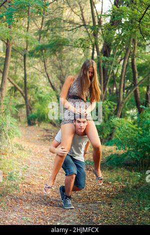 il giovane bel ragazzo con una ragazza minorenne si sta divertendo nella foresta. Ritratto di adolescenti adulti che camminano nel parco. La ragazza siede sulle spalle Foto Stock