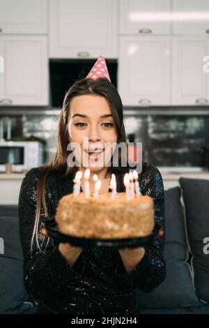 Giovane donna che soffia le candele su una torta di compleanno, una ragazza da sola in un cappello di festa soffia una candela, celebrazione a casa, felice concetto di compleanno Foto Stock