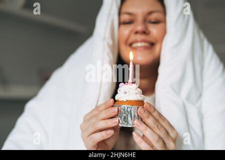 Giovane adulto felice bella donna quarant'anni più taglia corpo positivo in coperta bianca con cupcake festivo con candela in mani sul letto a casa pa Foto Stock