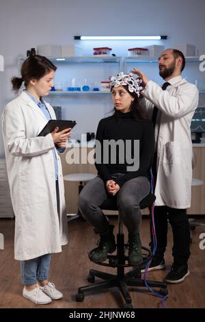 Medico di scienziato che spiega i sintomi di malattia neurologica a paziente di donna mentre ricercatore neurologo che regola la cuffia eeg durante l'esperimento di neuroscienza in laboratorio. Specialista che analizza l'attività cerebrale Foto Stock