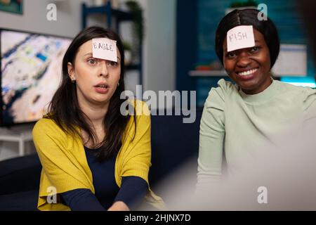 Ritratto di amici multietnici con note adesive sulla fronte giocare chi sono gioco godere di passare il tempo insieme appendere a casa in salotto. Gruppo di persone che si divertono durante la festa di wekeend Foto Stock