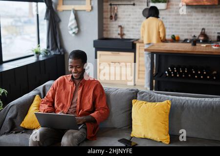 Ritratto del giovane afroamericano utilizzando un computer portatile mentre si rilassa sul divano in casa accogliente interno, spazio copia Foto Stock