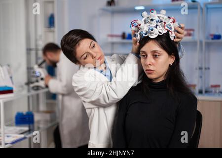 Donna scienziata che indossa la cuffia eeg sulla testa del paziente durante l'esperimento di neurologia in laboratorio medico. Medico neurologo analisi dell'evoluzione cerebrale monitoraggio dell'attività del sistema nervoso Foto Stock