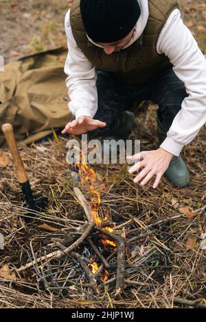 Colpo tagliato verticale dell'uomo di viaggiatore congelato che scalda le mani sopra il fuoco all'aperto nella giornata fredda overcast. Foto Stock