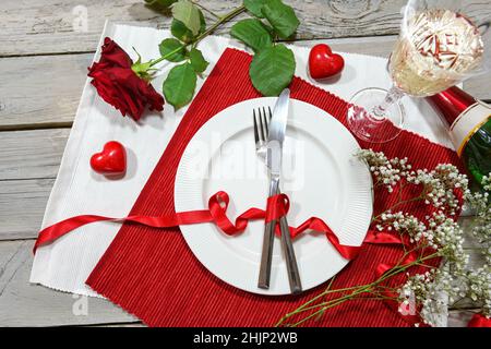 Romantico tavolo da pranzo con una rosa rossa, champagne e due cuori, piatto e posate su tovaglioli rossi e bianchi e un rustico tavolo in legno, alto angolo di vista Foto Stock