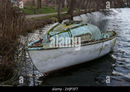 Piccola barca abbandonata in cabina in plastica rinforzata con fibra di vetro, cresciuta di muschio e lichene sulla riva di un lago in attesa di momenti migliori con Foto Stock