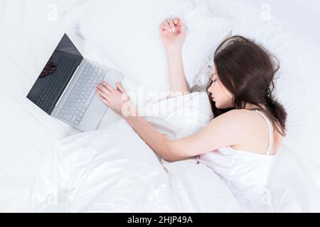 Buongiorno. La donna lavora a casa. Una ragazza di brunette si trova su un letto bianco nel suo pigiama, allungò la mano su un computer portatile. Foto Stock