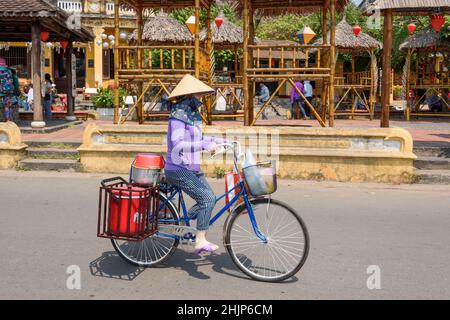 Una donna vietnamita che indossa un tradizionale cappello conico corre in bicicletta per le strade di Hoi An, provincia di Quang Nam, Vietnam centrale Foto Stock