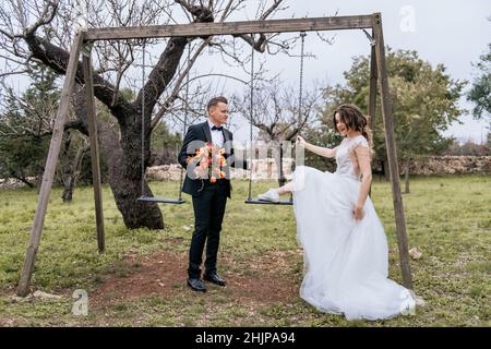 Felice coppia sorridente di stile che cammina in Toscana, Italia il giorno del matrimonio. SPOSI NOVELLI CON L'ALTALENA NEL PARCO. La sposa e lo sposo camminano giù il Foto Stock