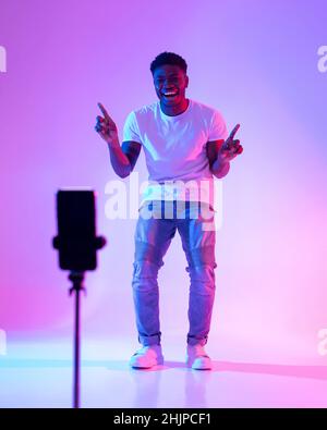 Giovane influencer afroamericano maschile che registra contenuti video, sorridendo alla webcam dello smartphone con luce al neon Foto Stock