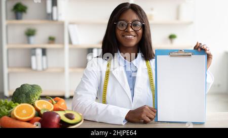 Medico americano africano nutrizionista tenendo appunti in bianco, mostrando mockup per il menu di nutrizione di destra, spazio di copia Foto Stock