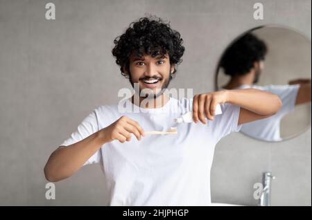 Concetto di pulizia dei denti. Giovane uomo indiano che applica dentifricio su spazzolino, spazzolando i denti in bagno la mattina Foto Stock