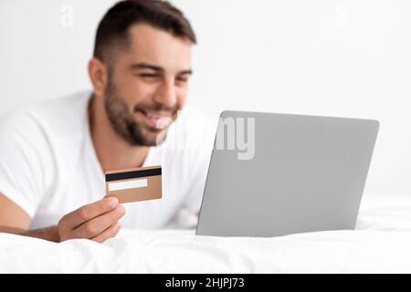 Felice caucasico attraente maschio millenario in bianco t-shirt acquistare online, ordini di acquisto su pc e mostra carta di credito Foto Stock