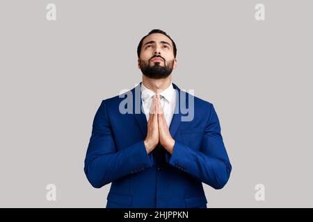 Ritratto di bel barbuto uomo che prega di cuore, sentendosi colpevole, supplicando il cielo implorando aiuto, indossando vestito di stile ufficiale. Studio interno girato isolato su sfondo grigio. Foto Stock