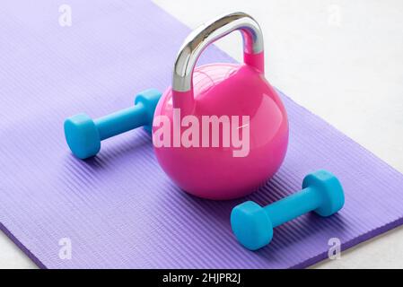 Primo piano di un peso luce blu su una stuoia viola yoga pilates sul pavimento di una palestra con un bollitore rosa Foto Stock