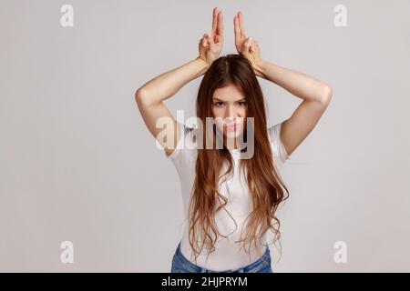 Ritratto di donna aggressiva bully con capelli scuri che mostrano corna toro gesto sopra la testa, accigliando come prima attacco, indossando T-shirt bianca. Studio interno girato isolato su sfondo grigio. Foto Stock