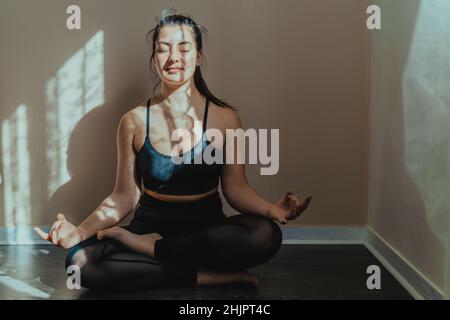 Giovane donna in posizione meditativa pratica yoga a casa - bella ragazza medita sukhasana postura relax - sano stile di vita corpo e mente c Foto Stock