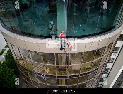 Operatore di alpinismo industriale che pulisce la finestra di un alto edificio mentre il collega si trova dietro il vetro. Pulitore uomo con attrezzatura di sicurezza durante il lavaggio della facciata grattacielo. Foto Stock
