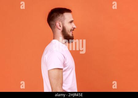 Ritratto laterale dell'uomo barbuto che guarda avanti e sorride, esprimendo emozioni positive, essendo di buon umore, indossando una T-shirt rosa. Studio interno girato isolato su sfondo arancione. Foto Stock