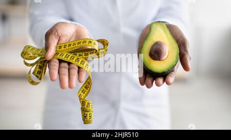 Concetto di stile di vita sano. Dietician femminile nero che tiene metà di avocado frutta e misurare nastro, closeup, panorama Foto Stock