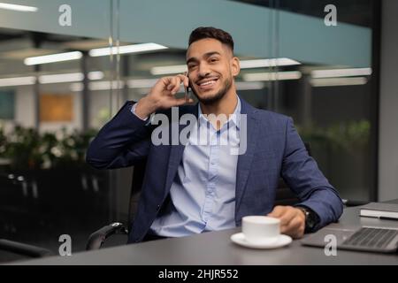 Allegro imprenditore arabo seduto al tavolo di lavoro, parlando al telefono Foto Stock