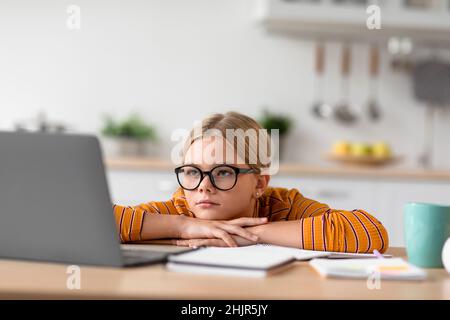 Stanco serio piuttosto europea adolescente pupilla in occhiali guarda il portatile, si siede a tavola Foto Stock