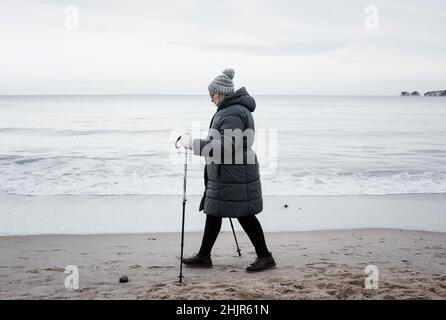 donna nel suo anni '70 a piedi con bastoni lungo la spiaggia in inverno Foto Stock