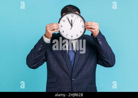 Sconosciuto uomo che indossa ufficiale abito di stile nascondendo faccia dietro la parete orologio display, sprecando il suo tempo, procrastinazione, organizzazione del tempo di lavoro. Studio interno girato isolato su sfondo blu. Foto Stock